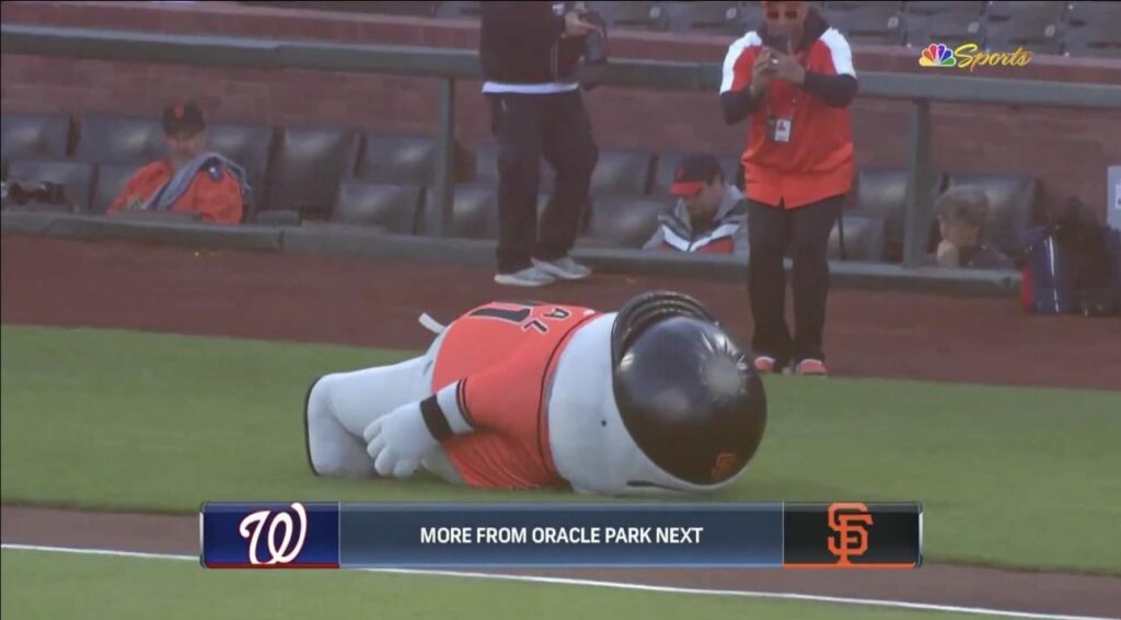 Screenshot of a TV broadcast showing a mascot lying face down on the ground